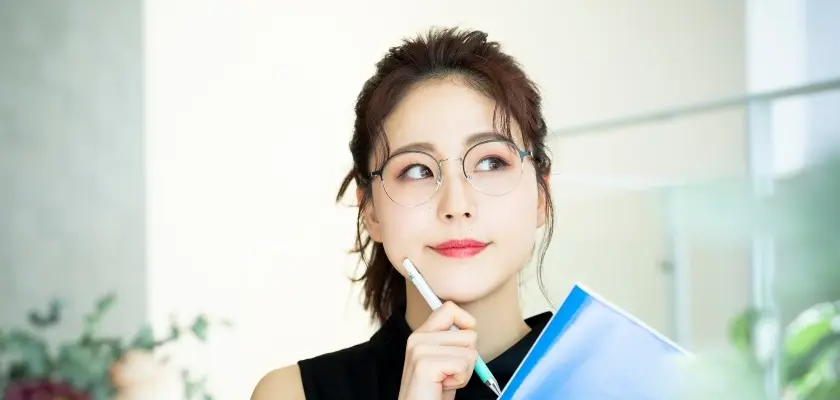 眼鏡をかけた女性が考える 眼鏡をかけた女性がペンを顎に当てて考えている様子。青いフォルダーを持ち、明るいオフィス環境にいる。