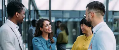 A group of four professionals having a friendly conversation in a modern office lobby, smiling and networking.