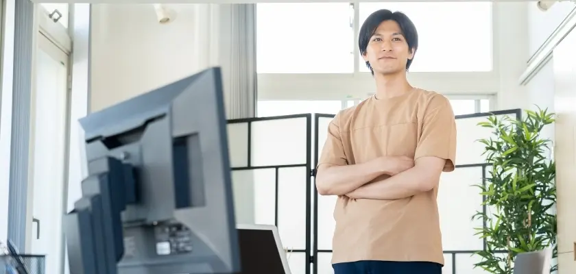 オフィスで立つビジネスマン 明るいオフィスで腕を組んで立つベージュのTシャツを着た若い男性。背景にはデスクトップモニターと観葉植物が見える。