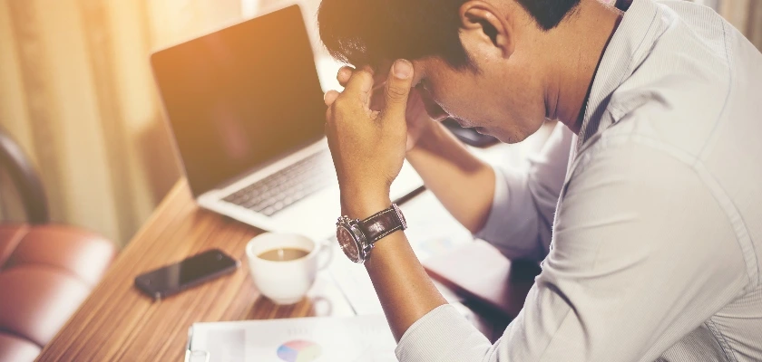 ノートパソコンとコーヒーのある机で頭を抱える男性の様子